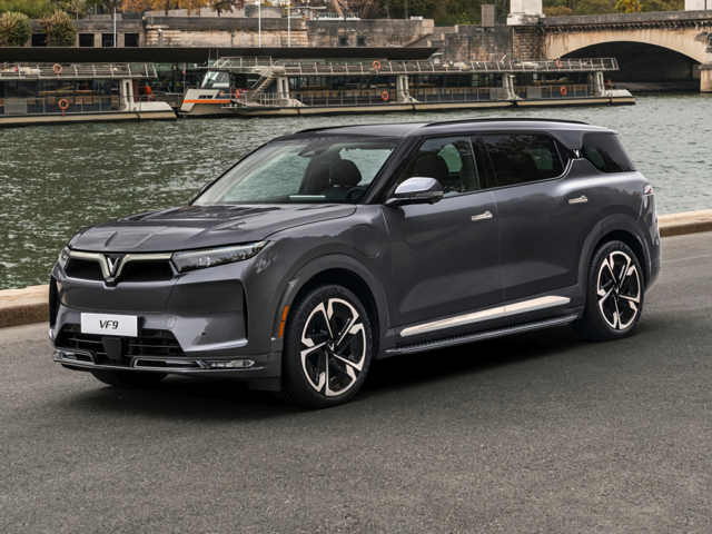Gray VinFast VF9 SUV parked on a waterfront roadway with a river, docked boats, and a bridge visible in the background