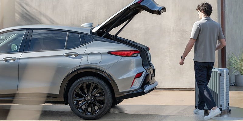 A person pulls a silver suitcase toward the open trunk of a gray SUV with black wheels