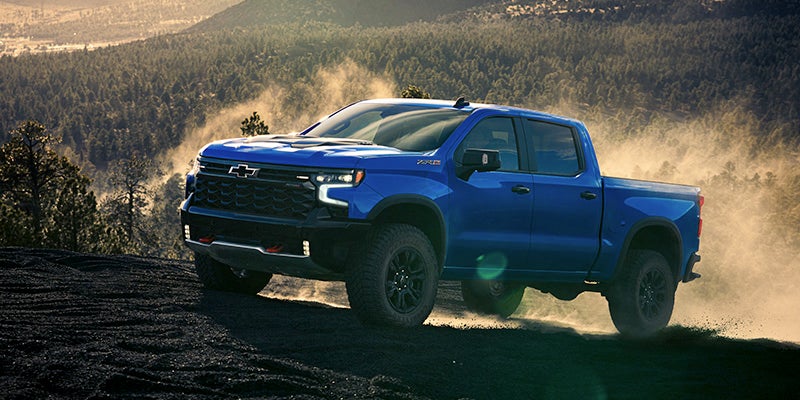 Blue Chevrolet Silverado ZR2 pickup truck off-roading on a dusty, rocky slope with a forested mountain landscape