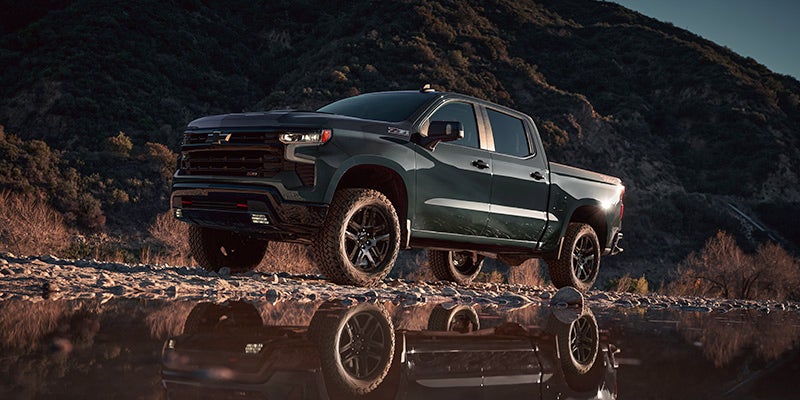 Dark green pickup truck with large tires, reflecting in water, against a dark mountain landscape under evening light