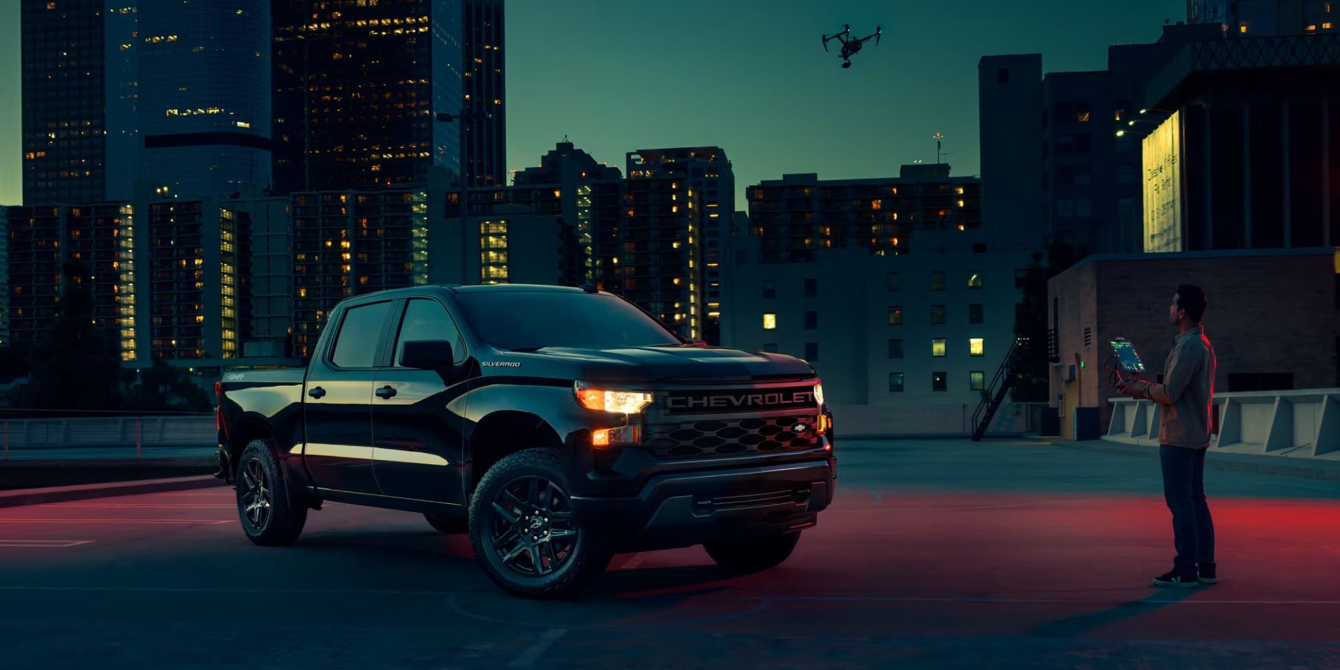 A black Chevrolet Silverado pickup truck parked on a rooftop lot at dusk with an operator flying a drone