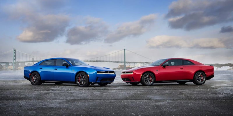 Blue 4-door and red 2-door Dodge Charger sedans with a bridge in the background under a cloudy sky.