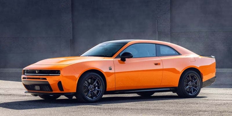 A bright orange Dodge Challenger or Charger muscle car, viewed from the side and front, parked on asphalt