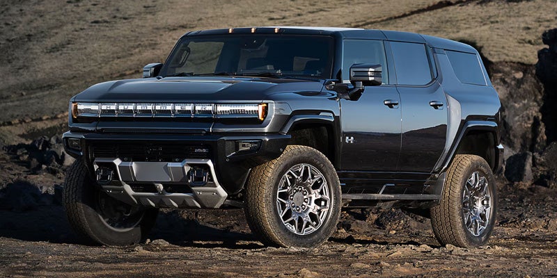 A dark gray Hummer EV SUV with bright LED headlights parked on a rocky, uneven dirt path