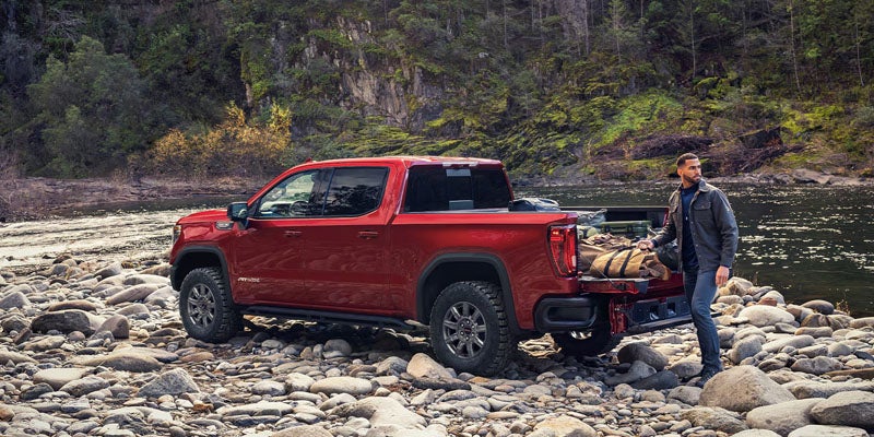A red pickup truck is backed up to a riverbank, tailbed open, with a man reaching into it to retrieve luggage