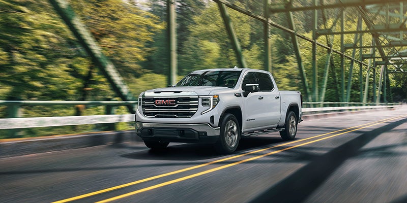 A silver GMC Sierra pickup truck drives across a green truss bridge, surrounded by trees and motion blur