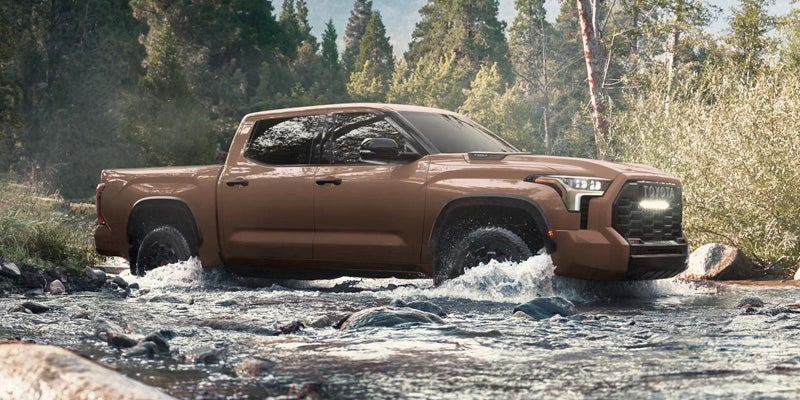 A brown Toyota Tundra truck driving through a rocky stream, surrounded by trees and lush foliage