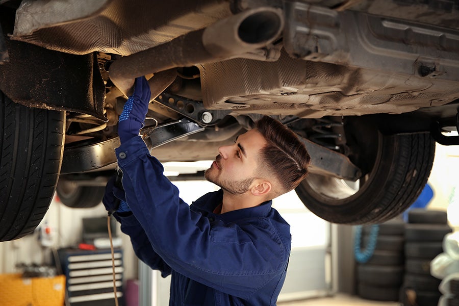 A car mechanic in blue coveralls inspecting the undercarriage and exhaust system of a vehicle raised on a lift