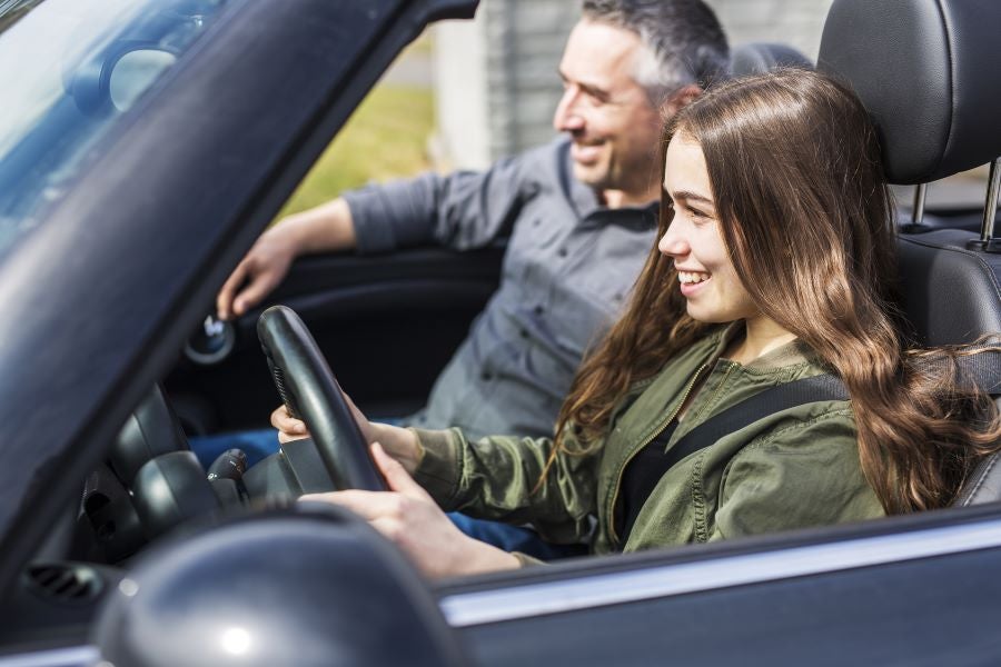 A smiling young woman driving a convertible car with her father sitting in the passenger seat